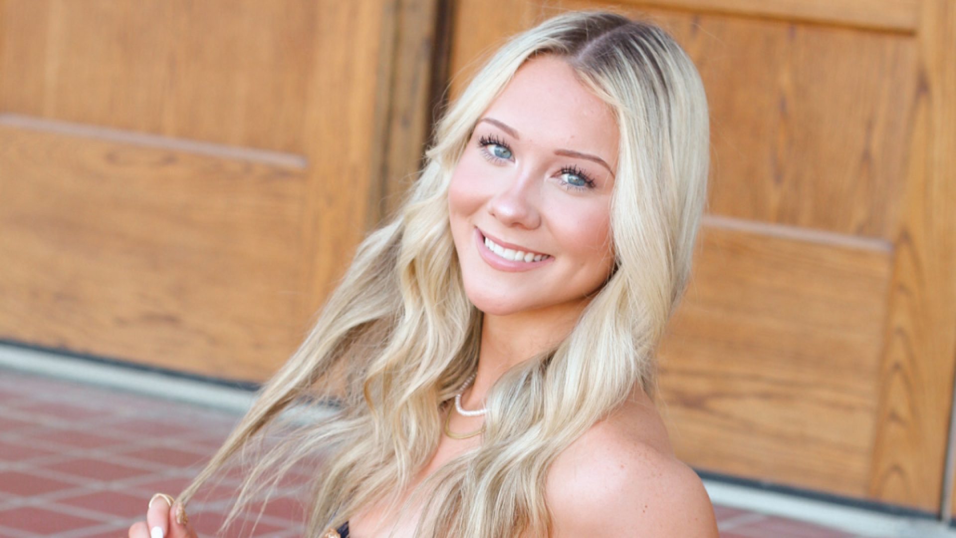 A young woman with long blonde hair smiles over her shoulder in front of wooden double doors.